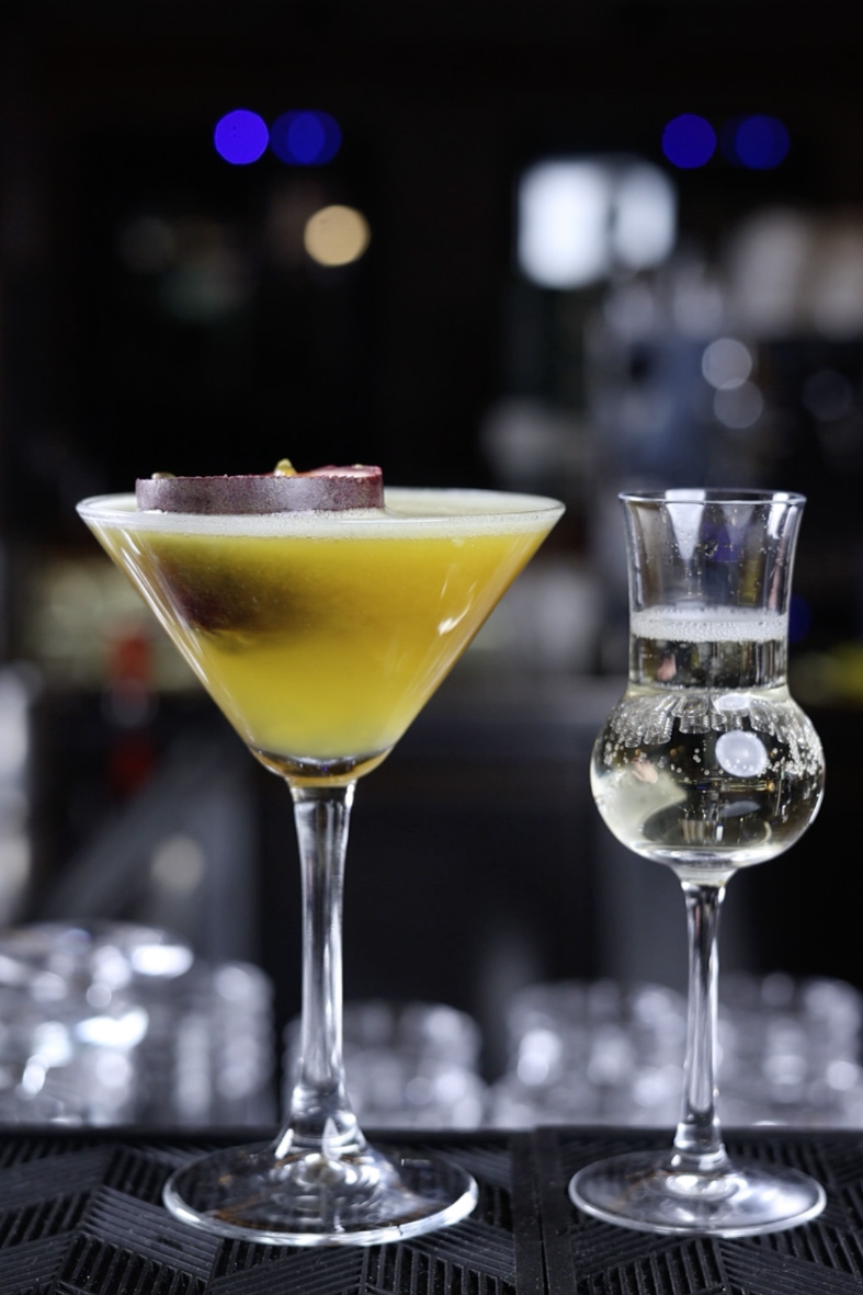 Pornstar-Martini in einem Glas mit einem Gläschen Prosecco daneben. Beides steht auf einer Bar.